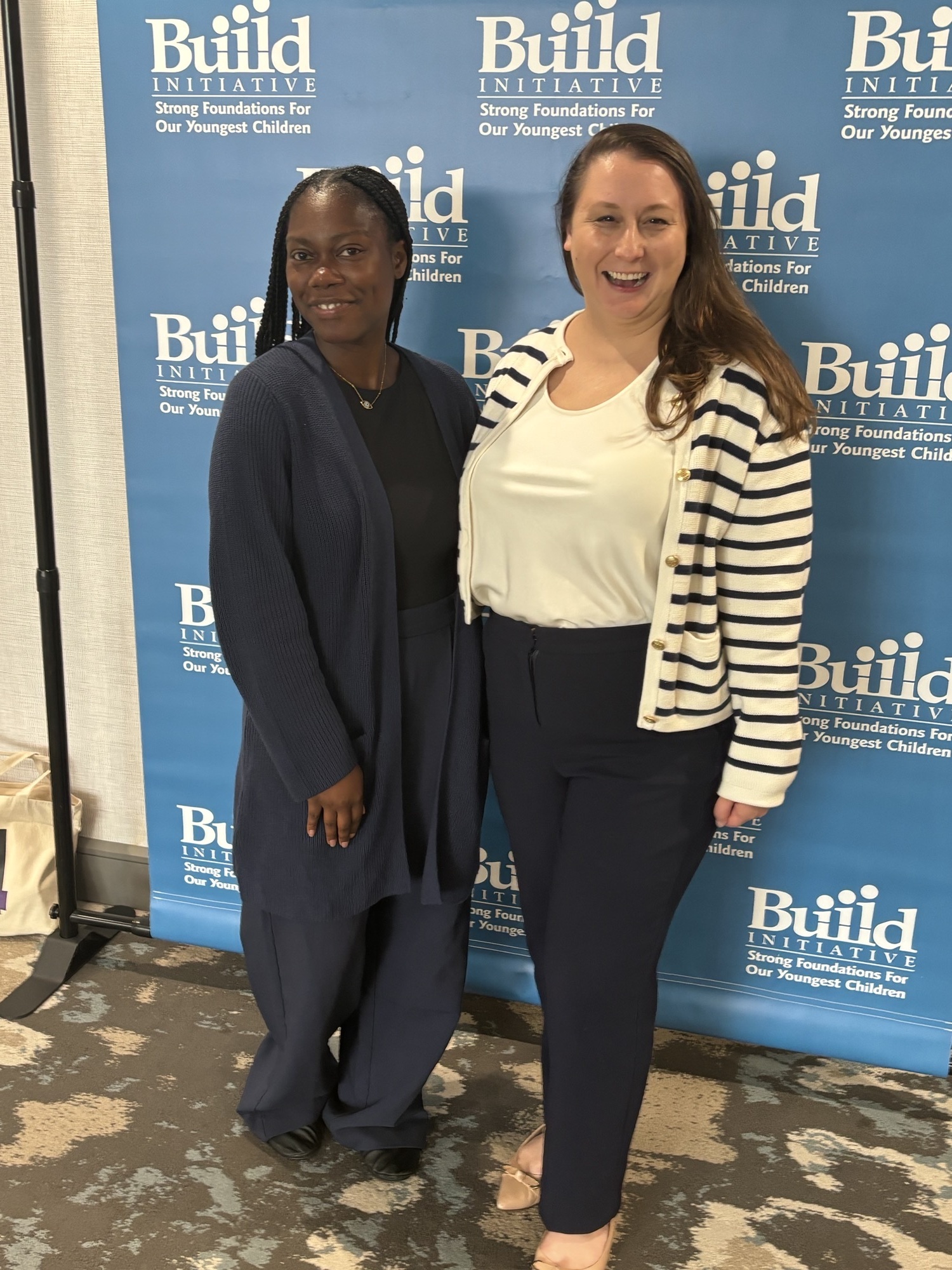 Two women pose in front of a step-and-repeat that says "Build Initiative" repeated, with a blue background and white text. The two women are smiling and posing after a presentation on child care policy at a conference.