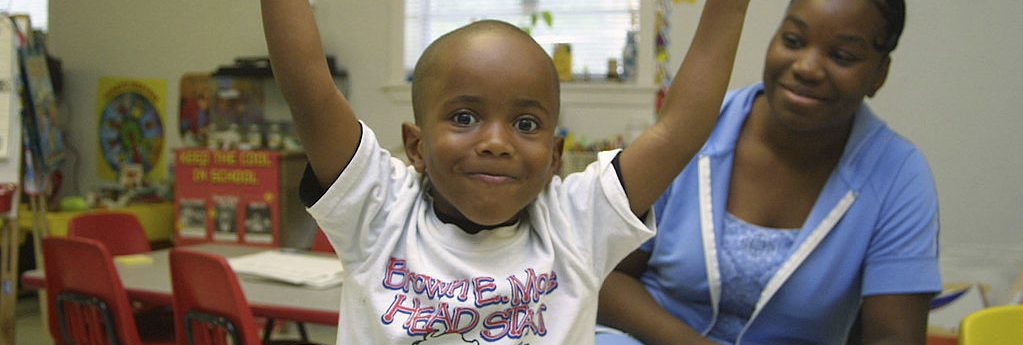 Child in Head Start program with childcare worker in background smiling and playing at daycare center